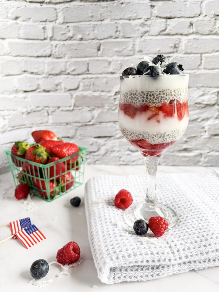 A parfait in a wine glass with layers of chia pudding, strawberries, and blueberries sits on a white towel. Fresh berries and an American flag pick are on the table; a green basket of strawberries is in the background against a white brick wall.