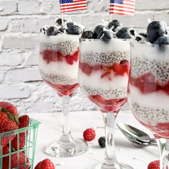 Three parfait glasses layered with chia pudding, yogurt, strawberries, raspberries, and blueberries are decorated with small American flags. A container of strawberries and scattered berries are on the white surface nearby.