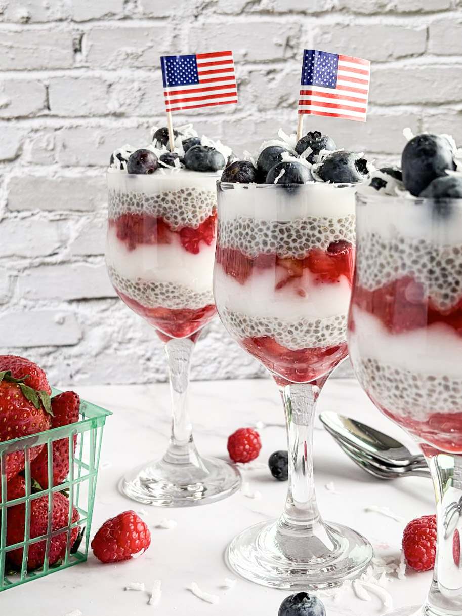 Three parfait glasses layered with chia pudding, yogurt, strawberries, raspberries, and blueberries are decorated with small American flags. A container of strawberries and scattered berries are on the white surface nearby.