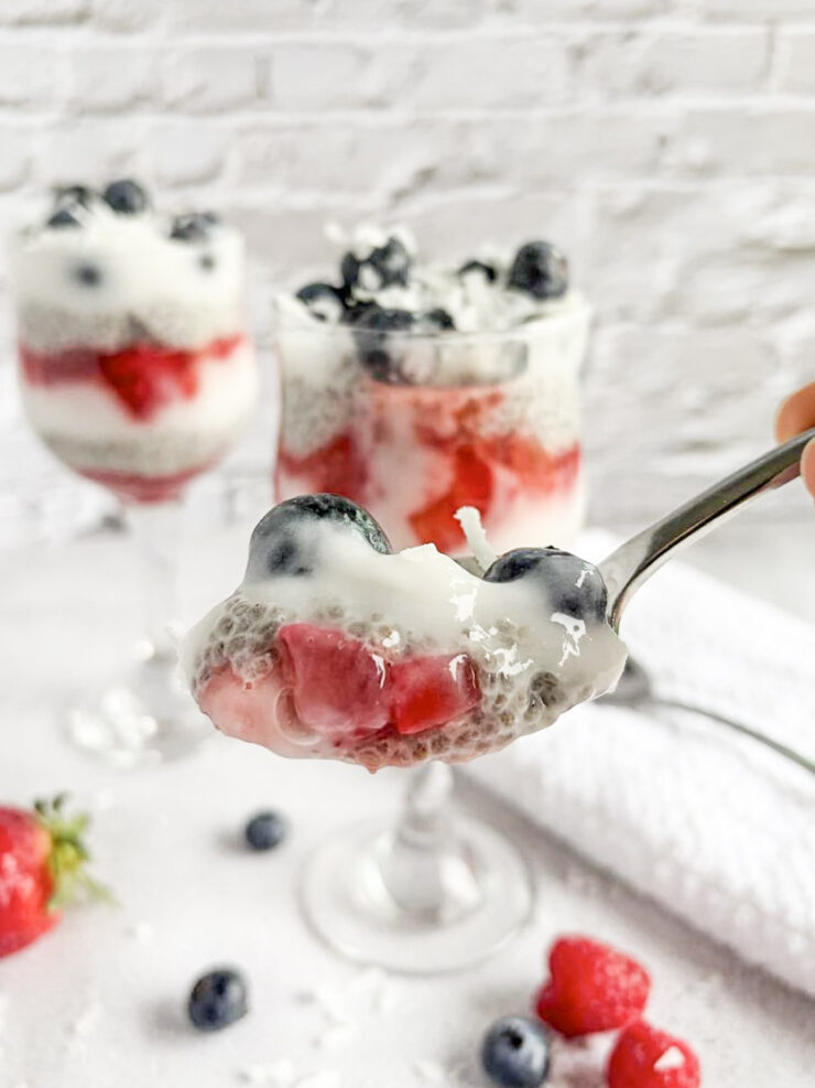 A close-up of a spoonful of chia pudding layered with strawberries, blueberries, and creamy topping, with two parfait glasses of the dessert and scattered berries in the background.