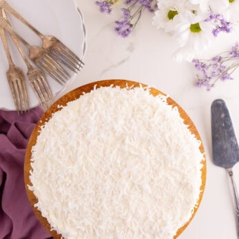 A Gluten-Free Dairy-Free Coconut Cake topped with shredded coconut on a wooden cake stand, surrounded by a cake server, forks on a plate, a purple napkin, and white and yellow flowers on a white surface.