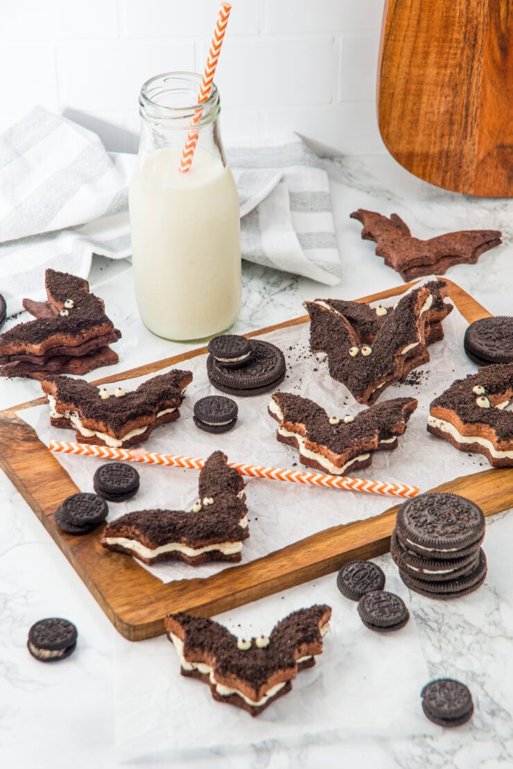 A wooden board with bat-shaped chocolate cookie sandwiches filled with cream, surrounded by Oreo cookies. A bottle of milk with an orange-striped straw and a striped napkin are in the background.