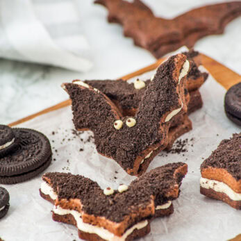 Chocolate sandwich cookies shaped like bats with cream filling and candy eyes are displayed on parchment paper, surrounded by round chocolate cookies and a striped orange straw.