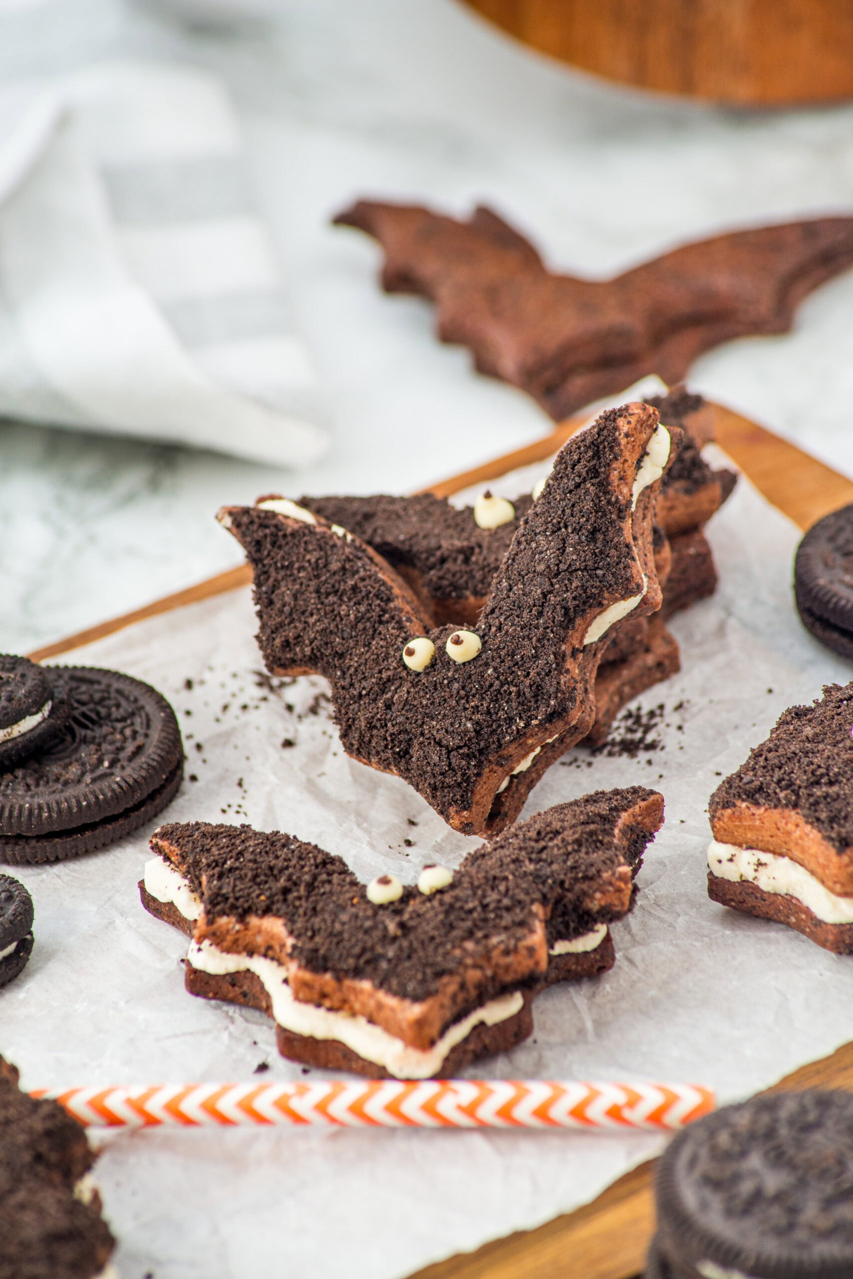 Chocolate sandwich cookies shaped like bats with cream filling and candy eyes are displayed on parchment paper, surrounded by round chocolate cookies and a striped orange straw.
