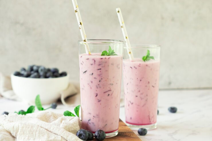 Two tall glasses filled with a pink blueberry smoothie, topped with mint leaves and striped straws. Blueberries and a white bowl of blueberries are in the background on a light surface.