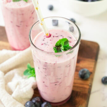 A glass of pink blueberry smoothie garnished with mint leaves and a straw, placed on a wooden board with fresh blueberries and mint leaves scattered around, and a bowl of blueberries in the background.