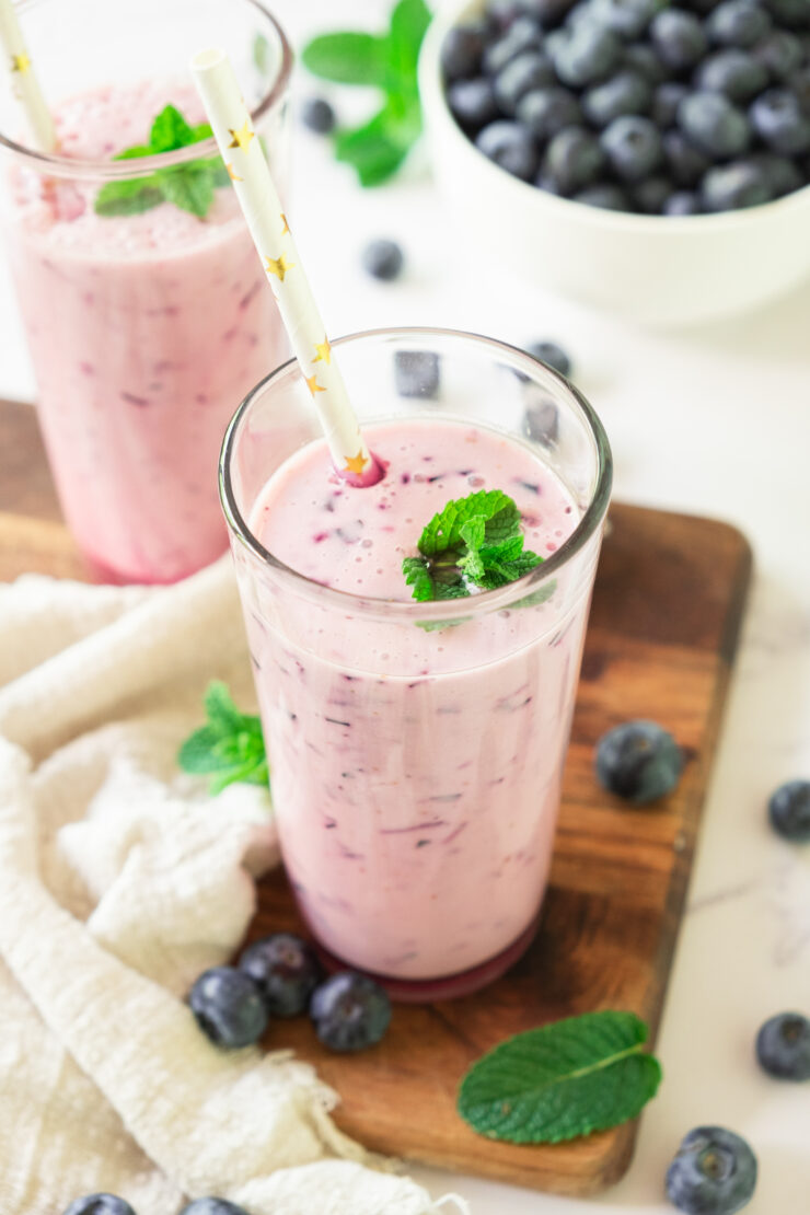 A glass of pink blueberry smoothie garnished with mint leaves and a straw, placed on a wooden board with fresh blueberries and mint leaves scattered around, and a bowl of blueberries in the background.