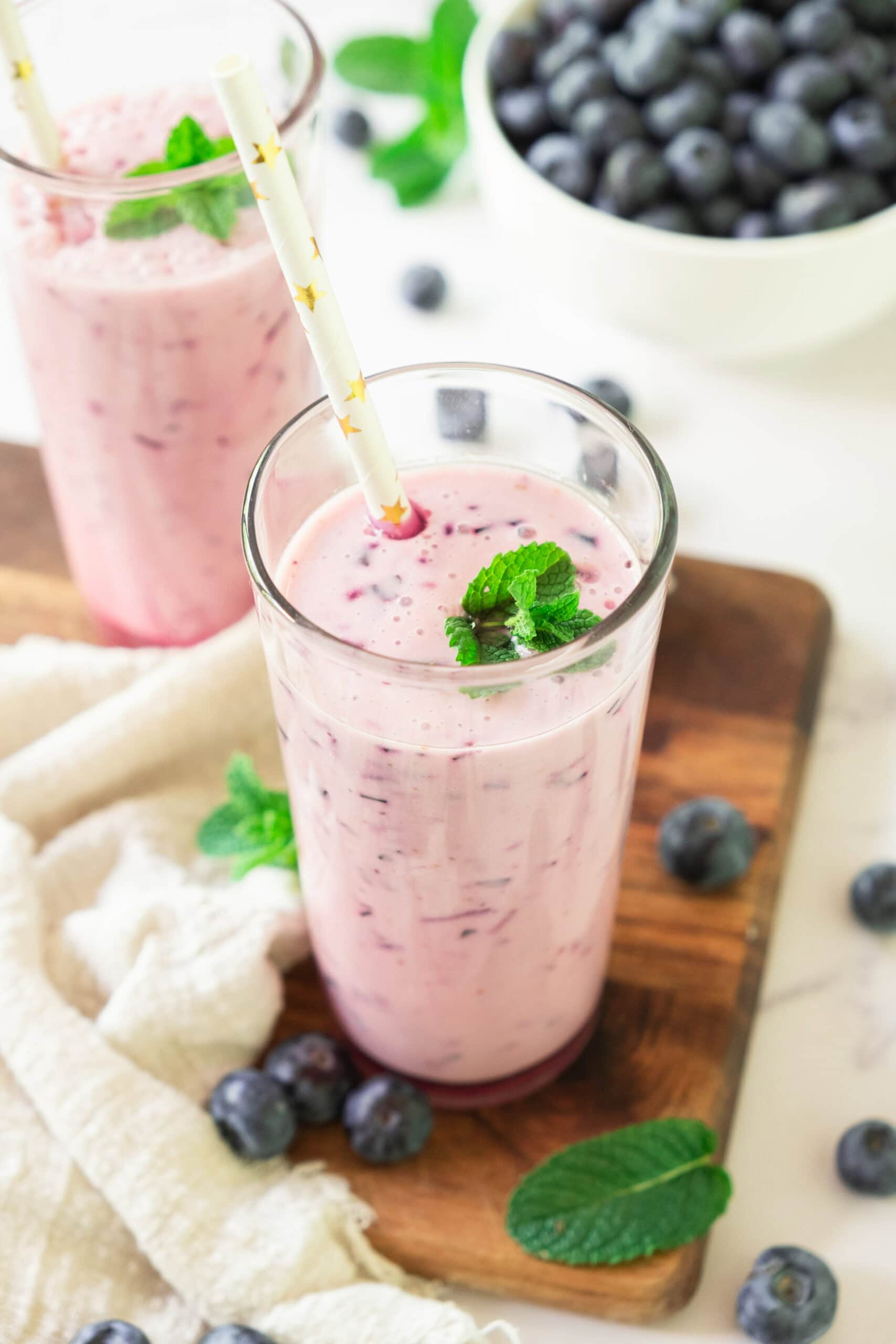 A glass of pink blueberry smoothie garnished with mint leaves and a straw, placed on a wooden board with fresh blueberries and mint leaves scattered around, and a bowl of blueberries in the background.