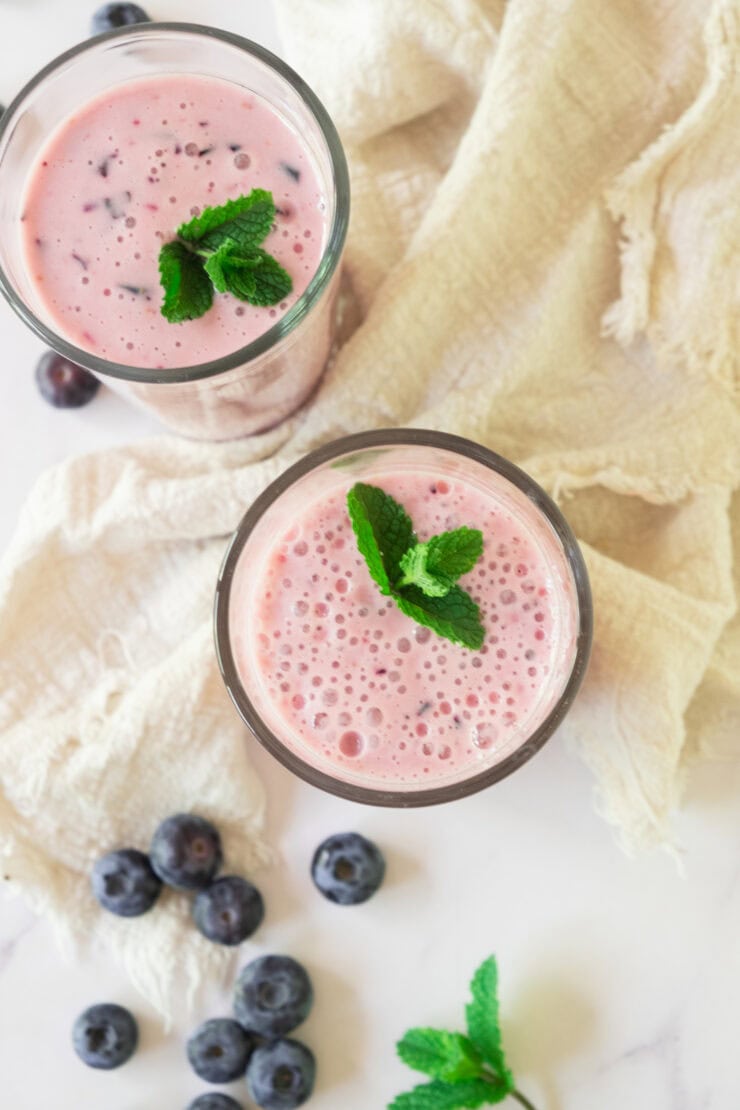 Two glasses of pink blueberry smoothie topped with fresh mint leaves, placed on a light-colored cloth with scattered blueberries and a sprig of mint on a white surface.