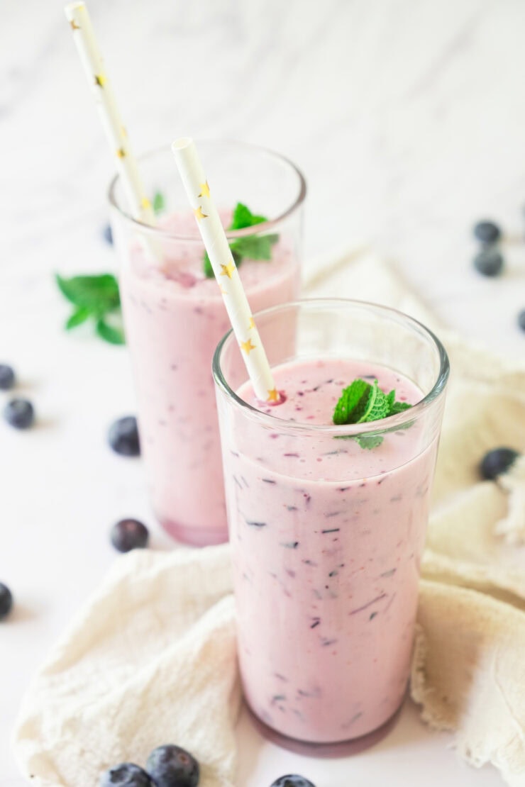 Two tall glasses of blueberry smoothie garnished with mint leaves and paper straws, surrounded by fresh blueberries and a light beige cloth on a white surface.