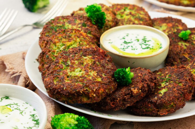 A plate of golden-brown vegetable fritters garnished with parsley and served with a bowl of creamy dipping sauce, with pieces of broccoli on the side.