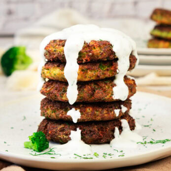 A stack of Broccoli Fritters (Gluten-Free)topped with creamy white sauce, garnished with chopped herbs, and served on a white plate with broccoli pieces.