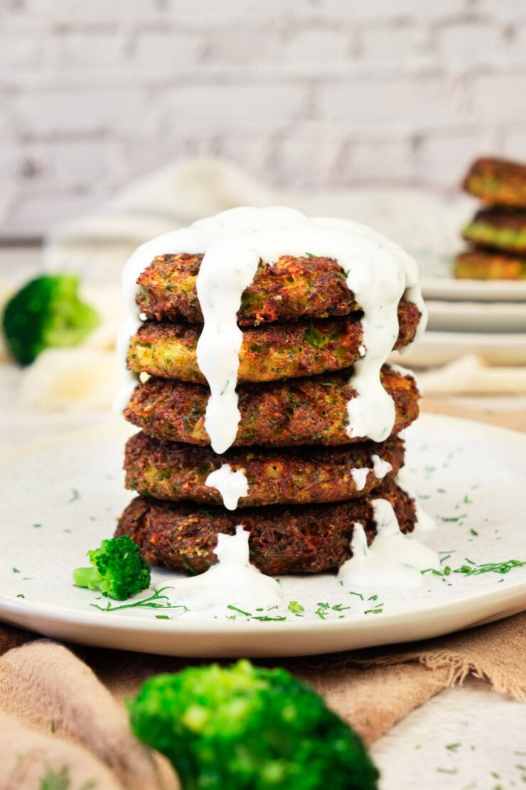 A stack of five golden-brown vegetable fritters topped with creamy white sauce, garnished with chopped herbs, and served on a white plate with broccoli pieces.