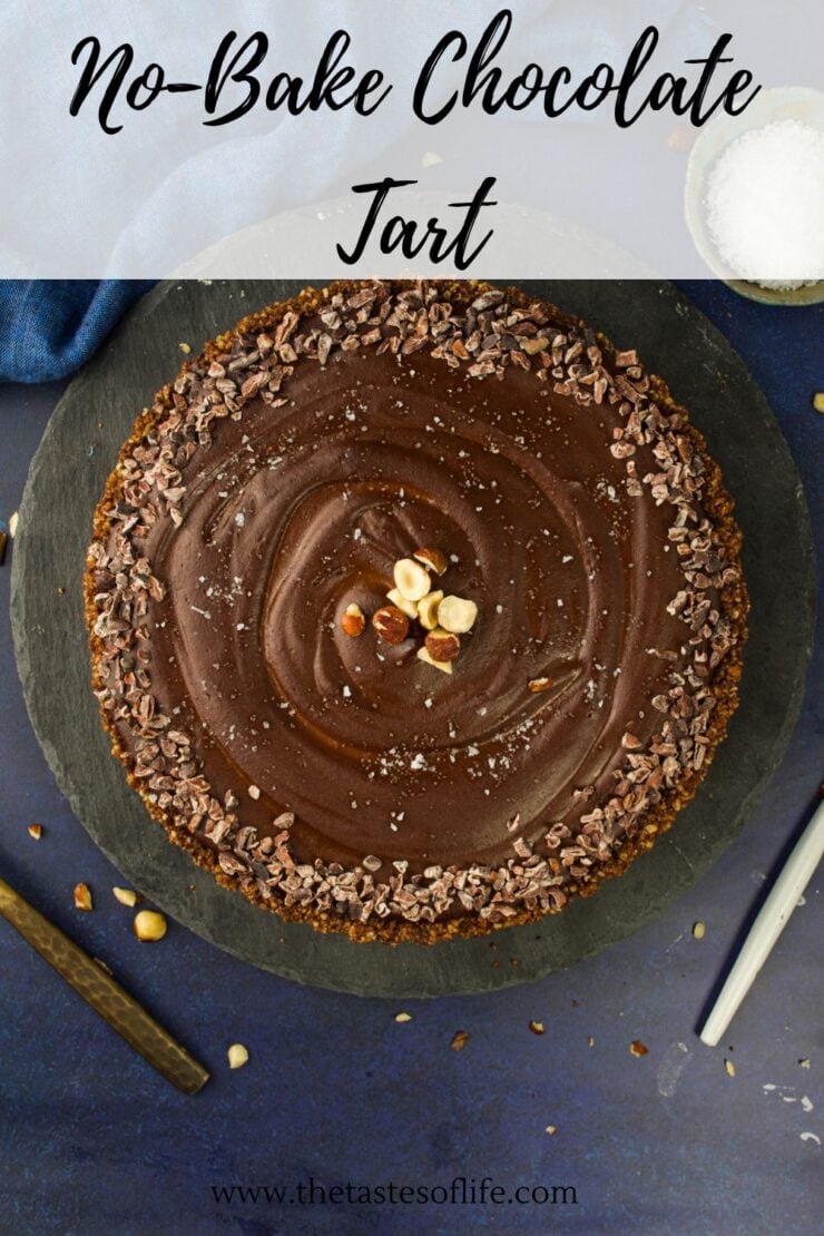 Top view of a no-bake chocolate tart garnished with cocoa nibs and hazelnuts on a black slate plate, with a pen, a white bowl of salt, and crumbs on a dark blue surface. Text at top: No-Bake Chocolate Tart.