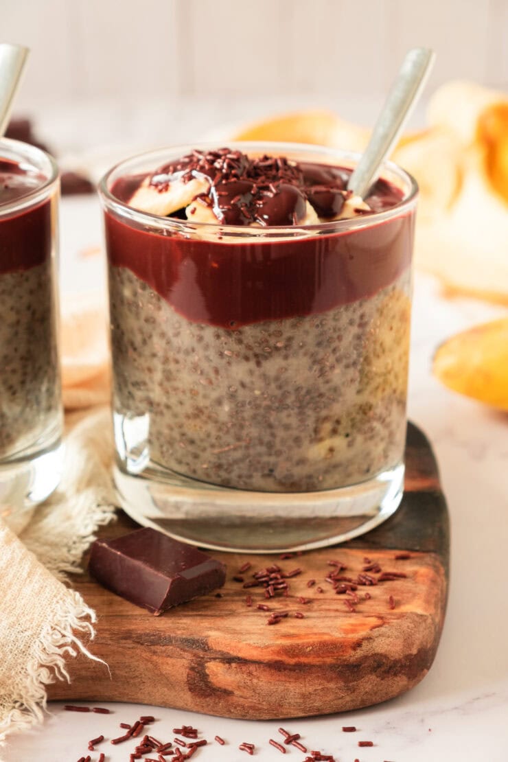 A glass filled with chia pudding topped with a thick layer of chocolate, chocolate sprinkles, and banana slices, placed on a wooden board with a piece of chocolate nearby. A spoon is in the glass.