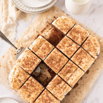 Creamy coconut and chocolate cake slice on parchment paper, milk in a pitcher, and a small chocolate cake piece on a plate.