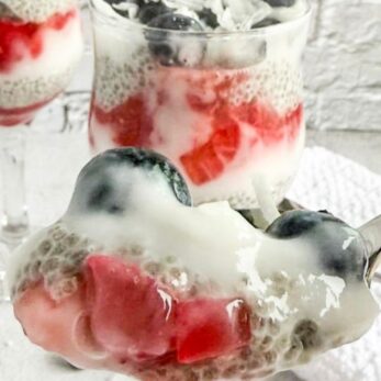 A close-up of chia pudding parfaits in glass cups, layered with blueberries, raspberries, and white pudding, with a spoonful in the foreground. The text reads, “Red, White & Blue Chia Pudding Parfait FROM SCRATCH.”.