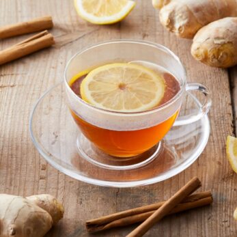 A glass cup of tea with a lemon slice, perfect for a Lymphatic Spring Cleanse, sits on a clear saucer atop a wooden table surrounded by fresh ginger roots, cinnamon sticks, and a lemon wedge.