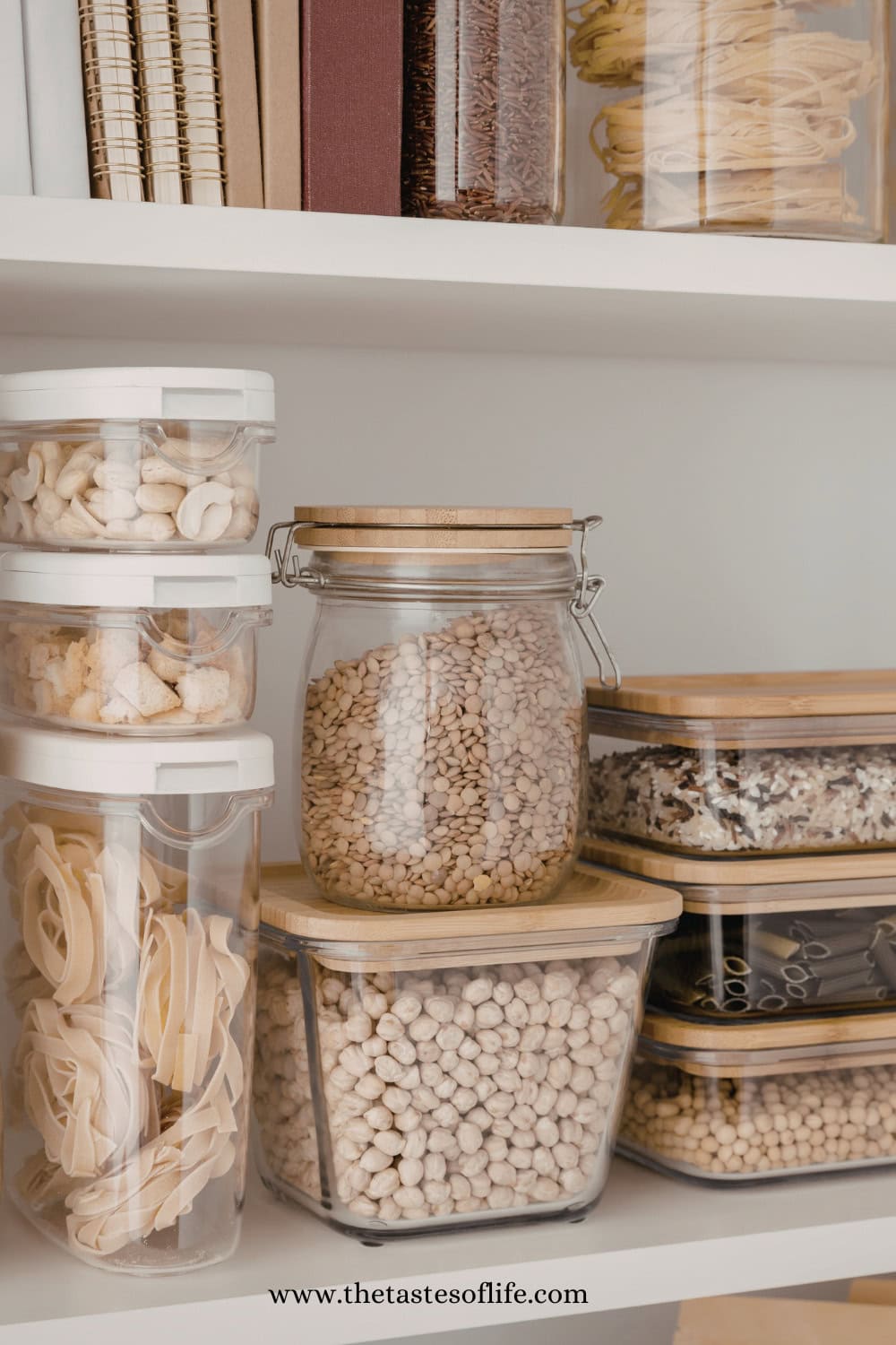 Clear containers filled with dried pasta, beans, chickpeas, grains, and nuts are neatly organized on white shelves, with some books on the top shelf. Jars have wooden lids, giving a tidy, minimalist pantry look.