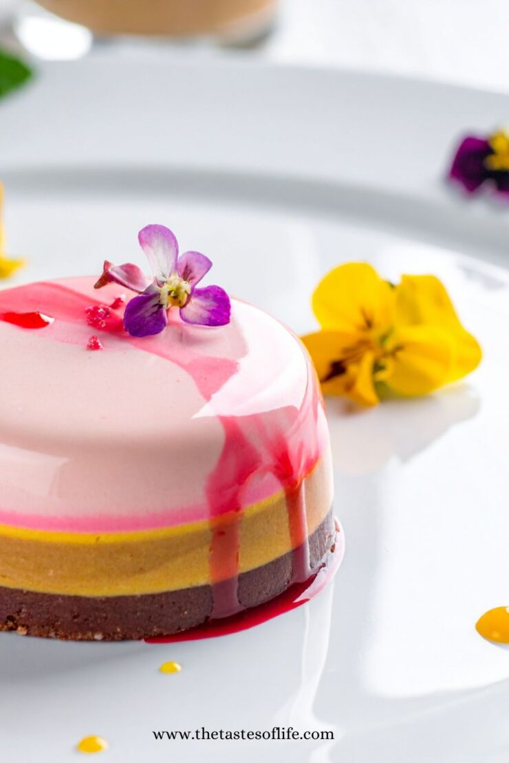 A layered mousse dessert with chocolate, yellow, and pink tiers, topped with a purple edible flower and red syrup, is plated with yellow and purple flowers on a white dish.