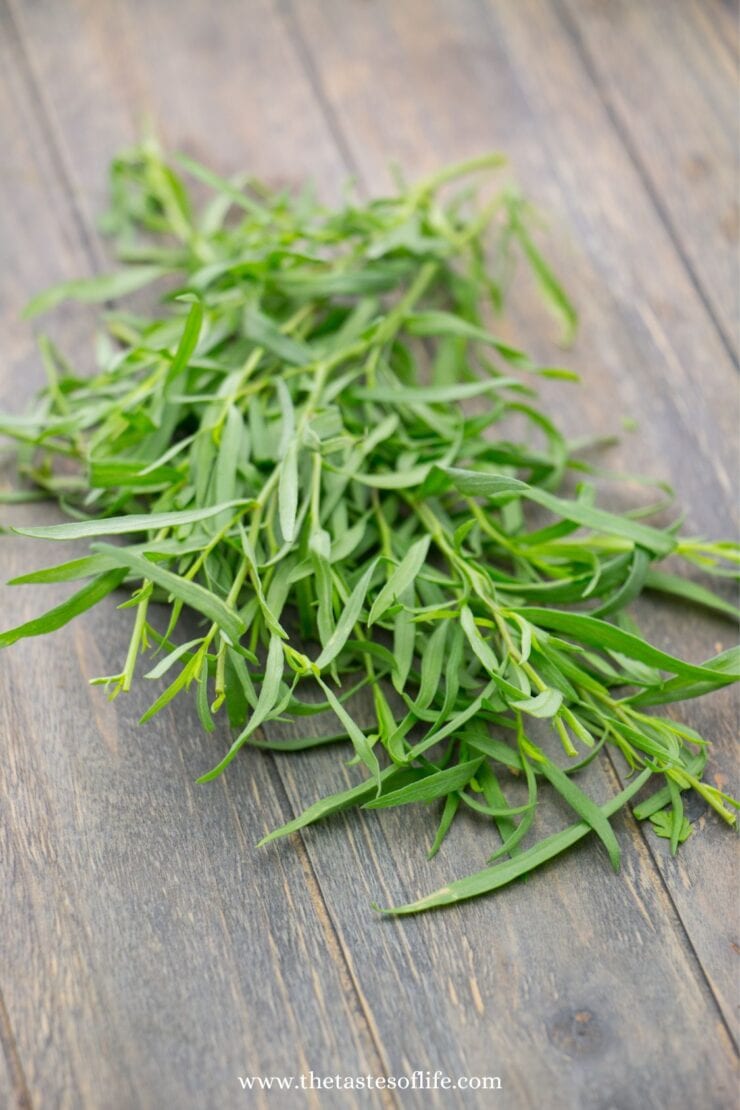 A bunch of fresh tarragon leaves rests on a rustic wooden surface, with thin, elongated green leaves spread out naturally.