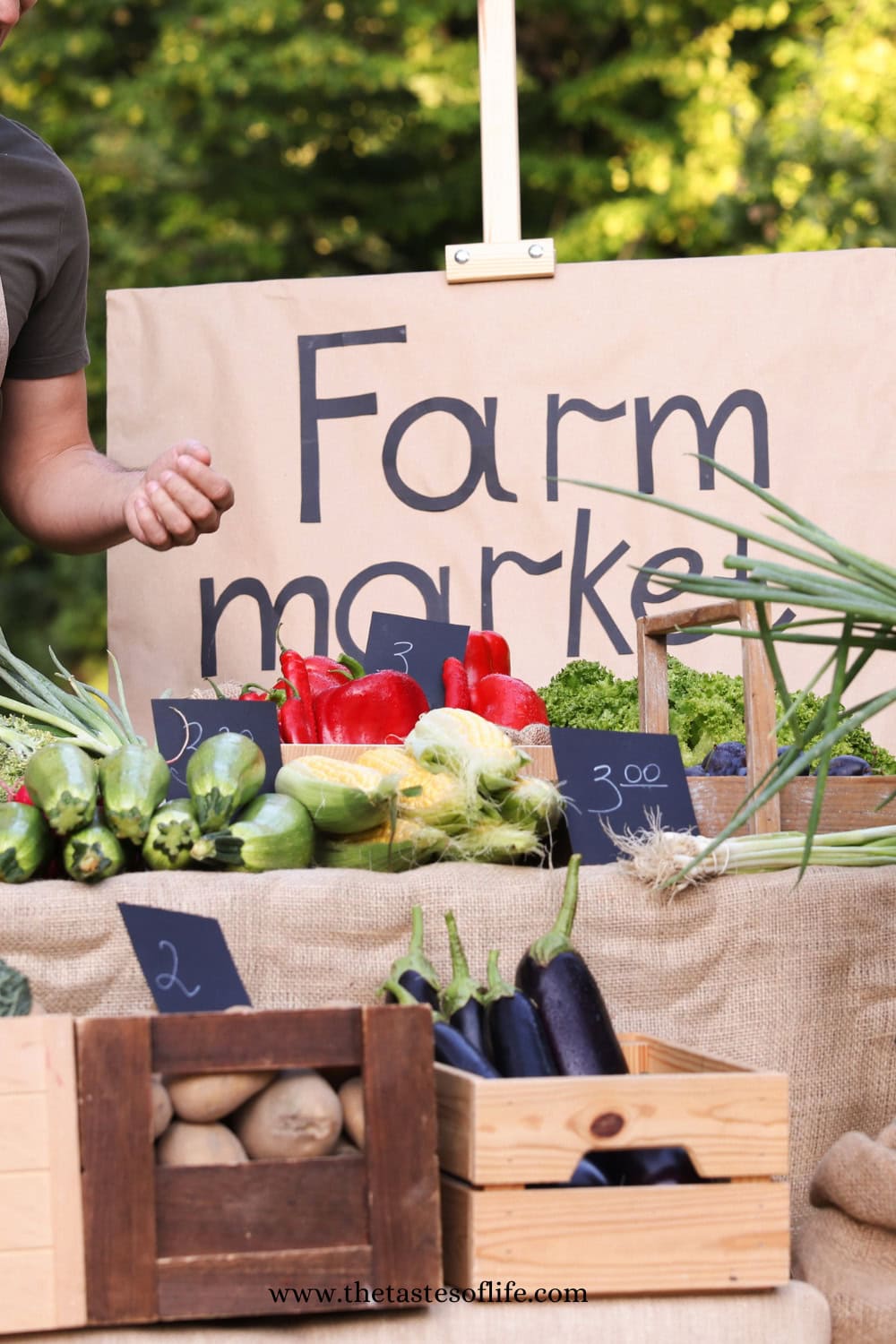 A farm market stand displays fresh vegetables including bell peppers, lettuce, zucchini, potatoes, and eggplants, with handwritten price signs on chalkboards. A person&rsquo;s arm is visible next to a large Farm market sign.