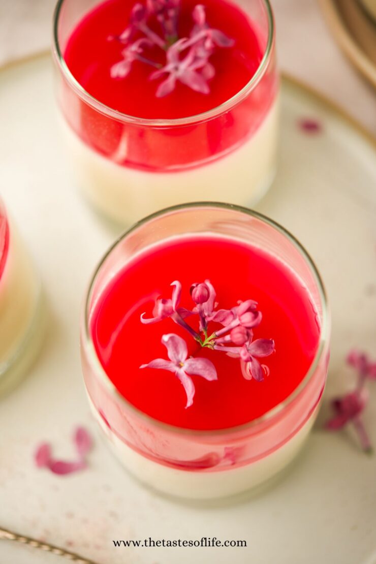A glass dessert with creamy white panna cotta topped with bright red jelly and pink edible flowers, placed on a light plate with flower petals around.
