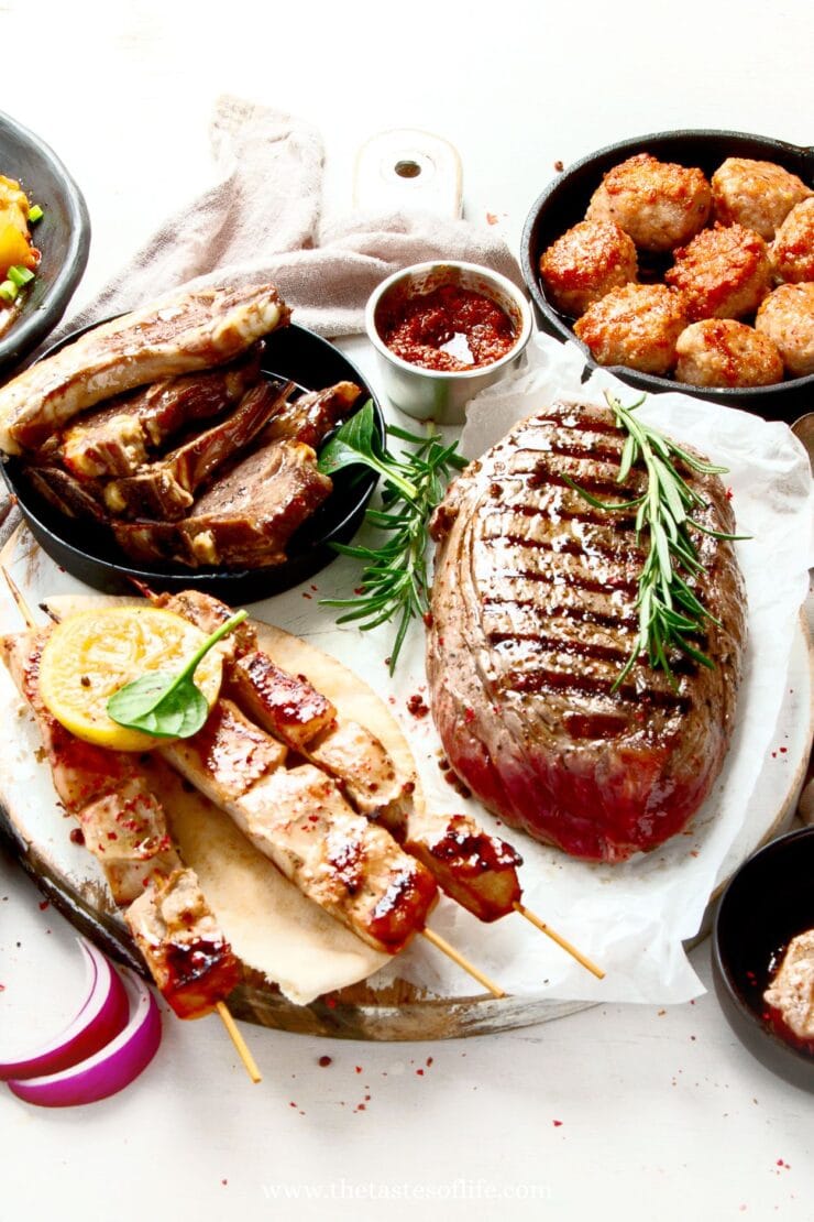 A variety of grilled meats including a large seasoned roast, lamb chops, meatballs, and skewered kebabs with lemon, arranged on plates with herbs and sauce on a white table.
