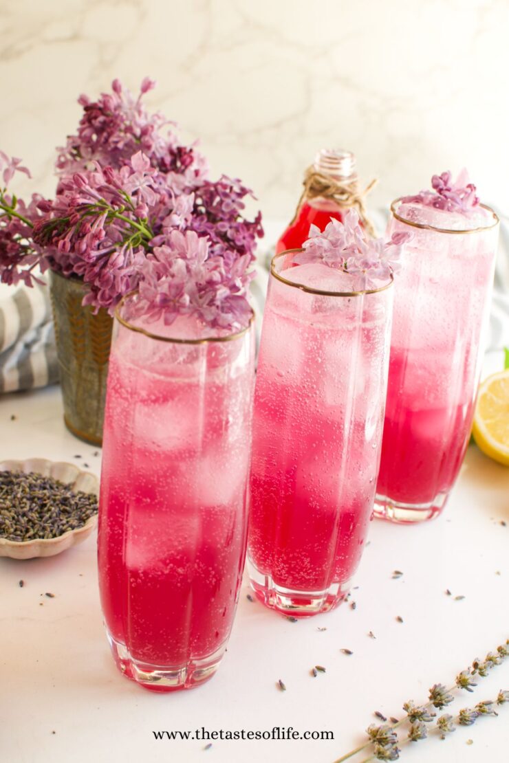 Three tall glasses of pink sparkling lemonade garnished with lilac flowers, with a vase of lilacs and a sliced lemon in the background. The setup is bright and refreshing, evoking a spring or summer feeling.