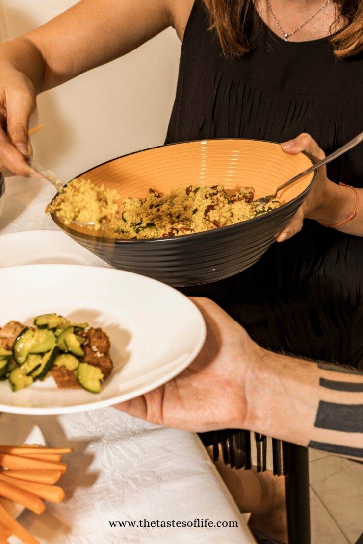 A person in a black sleeveless top serves a portion of couscous from a large bowl onto a dinner plate held by another person. Sliced carrots are visible on the table.