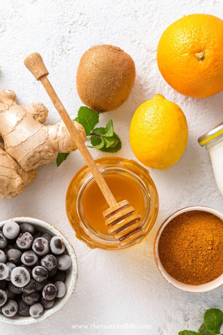 A top view of fresh ginger, a kiwi, an orange, a lemon, a jar of honey with a honey dipper, a bowl of frozen blueberries, a bowl of turmeric powder, and a jar of yogurt on a white surface.