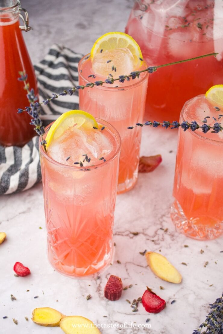 Three tall glasses of pink lemonade with ice, garnished with lemon slices and sprigs of lavender, sit on a marble surface with scattered dried fruits and ginger. A striped cloth and a pitcher of pink lemonade are in the background.