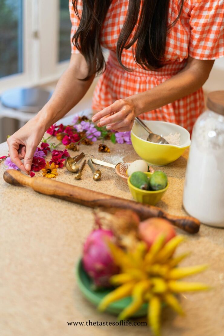 A woman in an orange-checkered dress arranges colorful flowers on a kitchen counter with baking tools, flour, limes, and tropical fruit nearby. The website thetastesoflife.com is visible at the bottom of the image.
