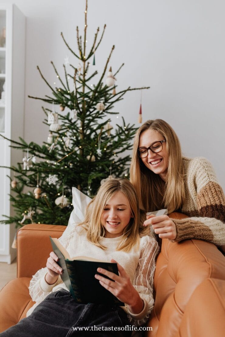 A woman and a girl sit on a brown sofa by a decorated Christmas tree. The girl is reading a book and smiling, while the woman, also smiling, holds a cup and looks at the book. The atmosphere is cozy and festive.