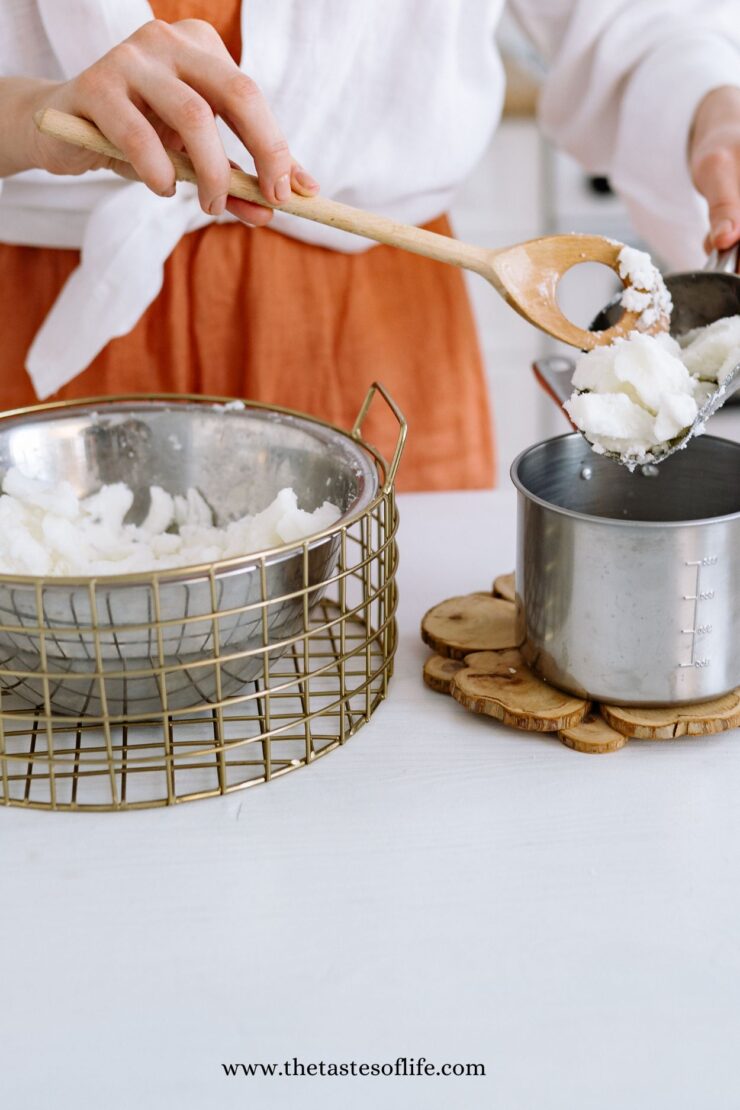 A person scooping coconut oil from a metal wire basket into a measuring cup using a wooden spoon, with a white background and the website www.thetastesoflife.com visible at the bottom.