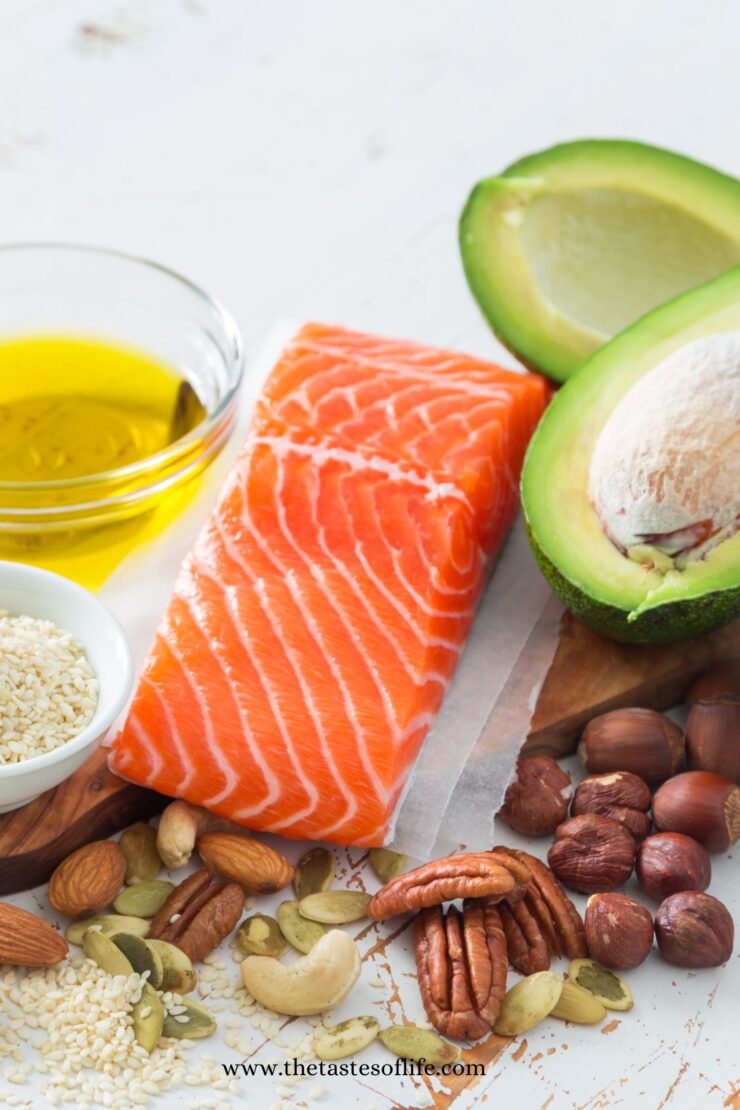 A fresh salmon fillet, halved avocado, bowl of olive oil, mixed nuts, and seeds are arranged on a white surface. The foods represent healthy sources of fats.