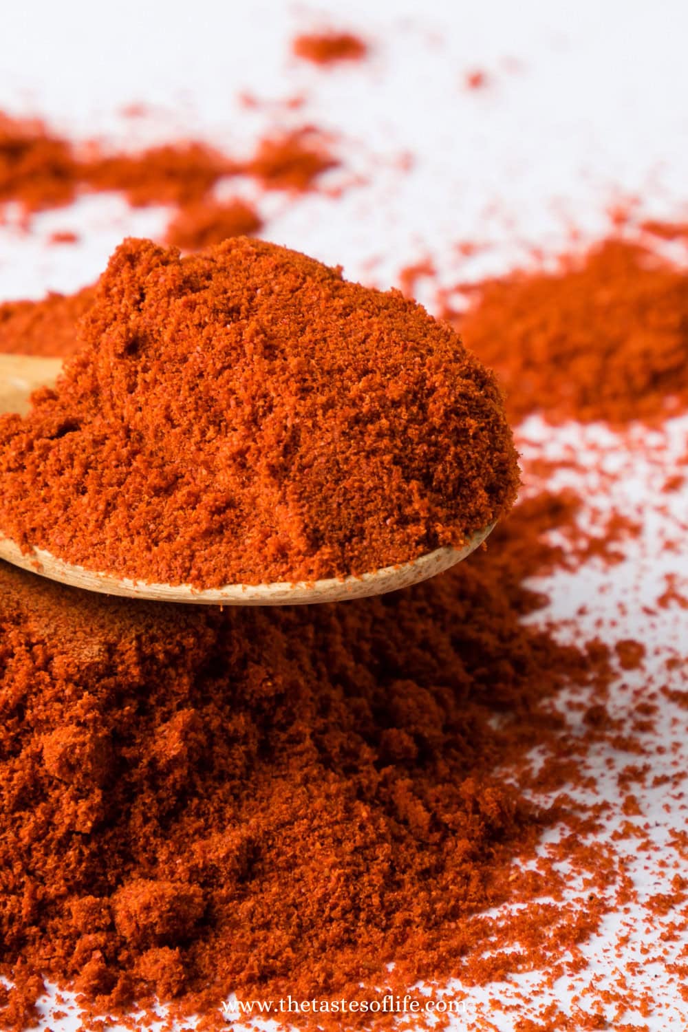 A wooden spoon holding a heap of vibrant red paprika powder rests on a pile of more paprika, with scattered powder visible on a white surface in the background.