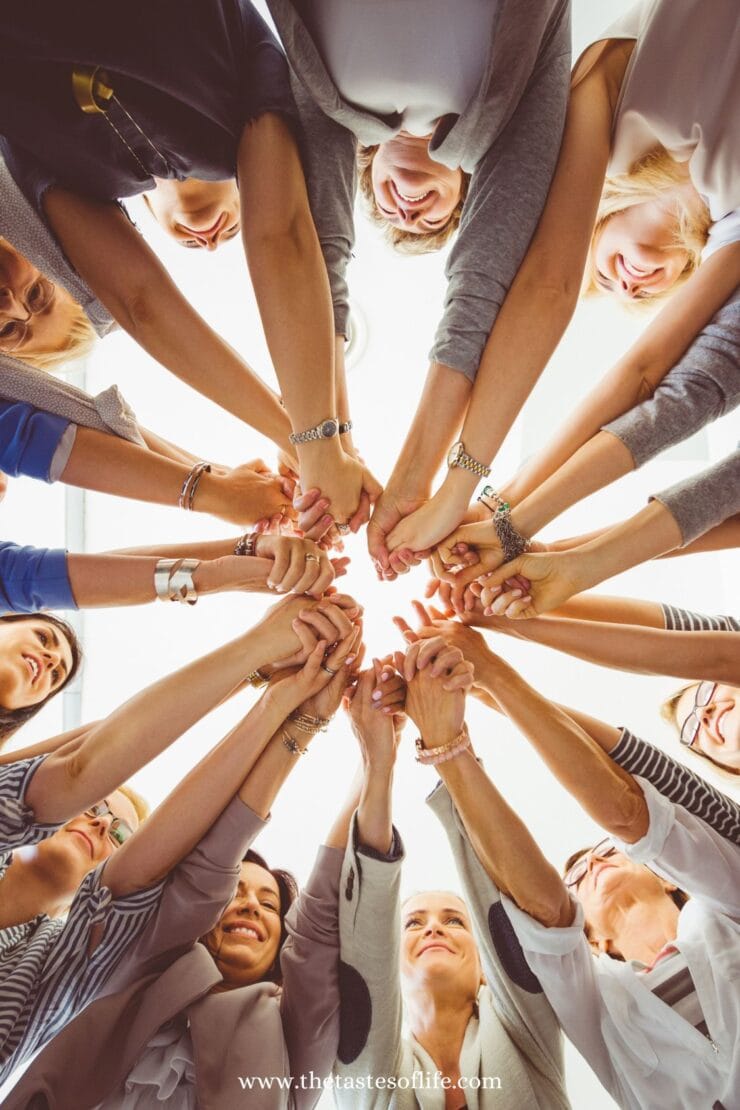 A group of people standing in a circle reach their hands toward the center, joining together in a show of unity and teamwork, viewed from below against a bright background.