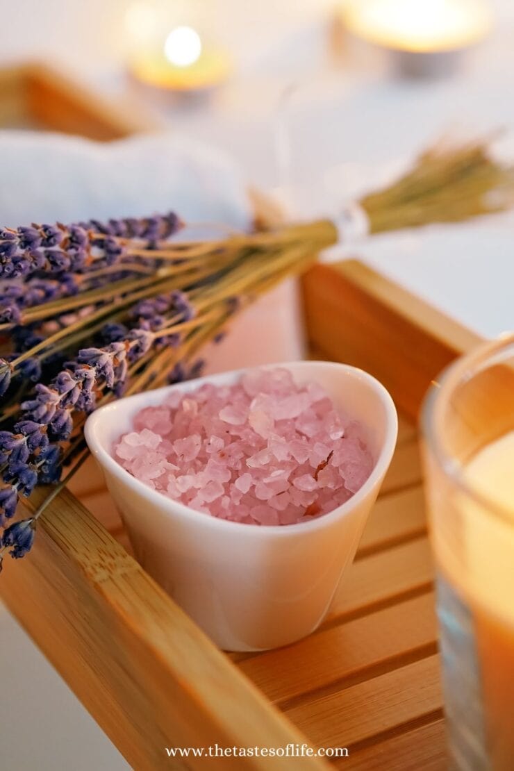 A white bowl filled with pink bath salts sits on a wooden tray next to a bunch of dried lavender, with candles glowing softly in the background.