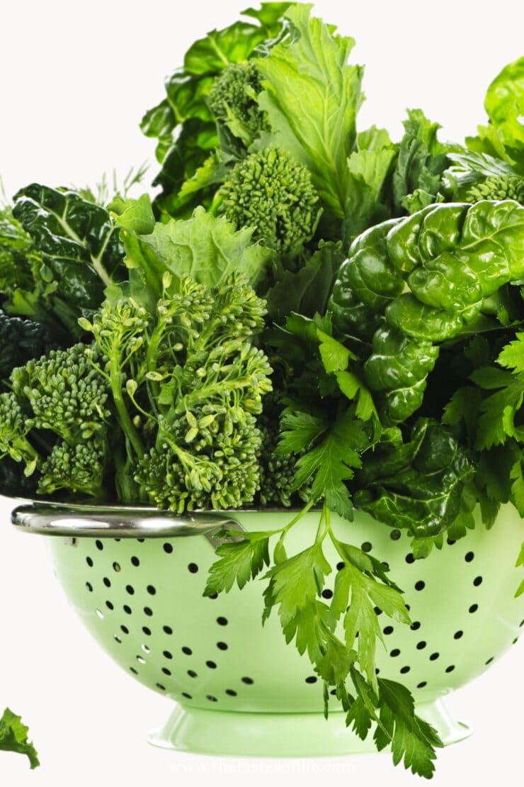 A green colander filled with fresh leafy greens, including broccoli, parsley, Swiss chard, and lettuce, against a white background.