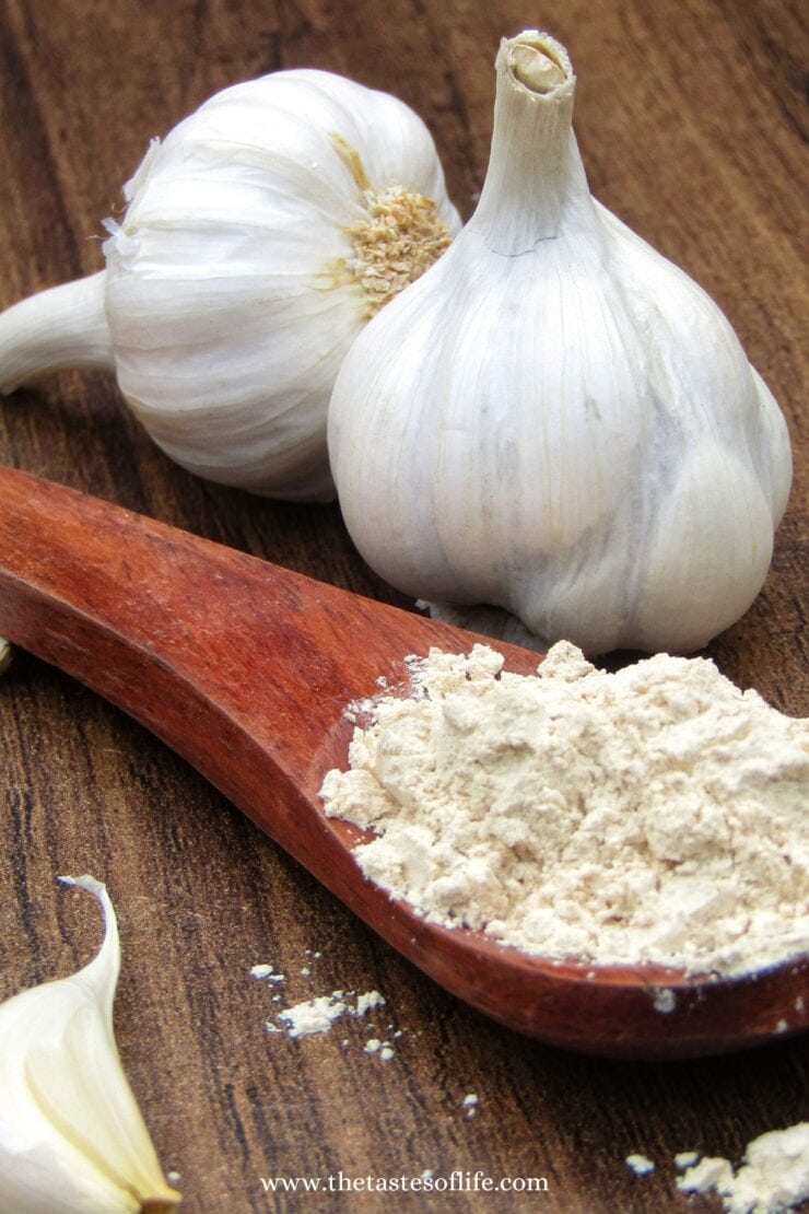 Two whole garlic bulbs and a garlic clove sit on a wooden surface next to a wooden spoon filled with garlic powder.