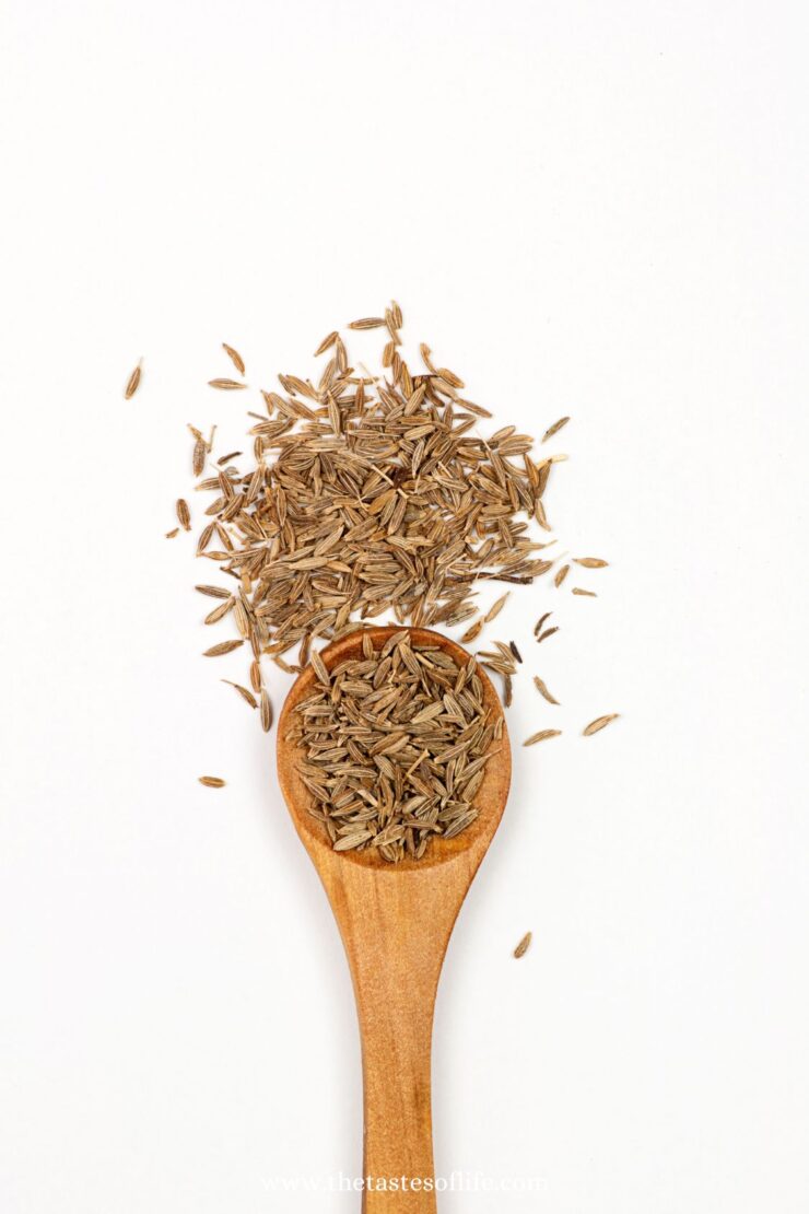 A wooden spoon filled with brown cumin seeds rests on a white surface, with more cumin seeds scattered around it.