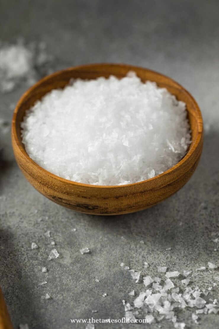 A small wooden bowl filled with coarse, flaky sea salt sits on a gray surface, with some salt crystals scattered around the bowl.