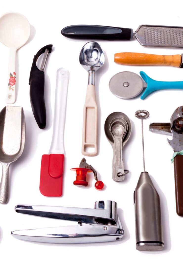 A variety of kitchen utensils arranged on a white background, including a scoop, peeler, spatula, ice cream scoop, pizza cutter, can opener, grater, whisk, and garlic press.