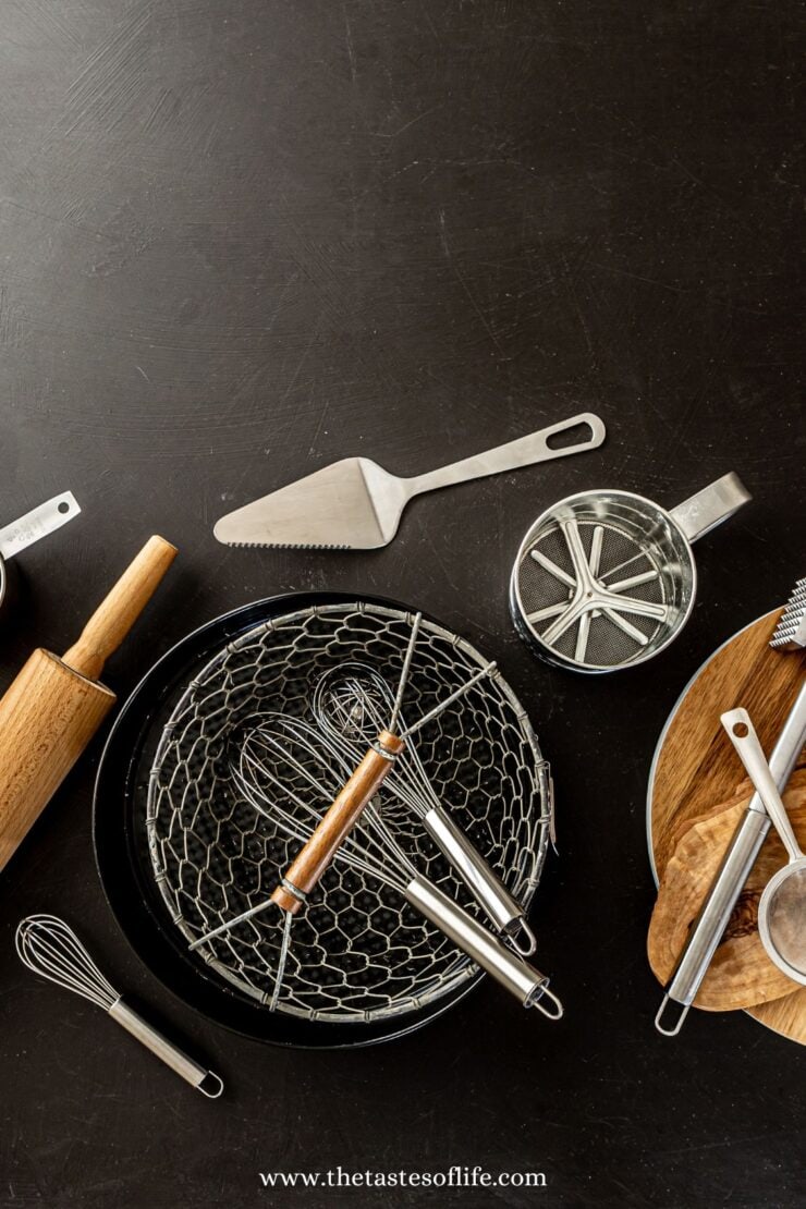 A variety of baking and cooking utensils, including a whisk, rolling pin, pastry cutter, sifter, and mesh trays, are arranged on a dark countertop with a wooden board.
