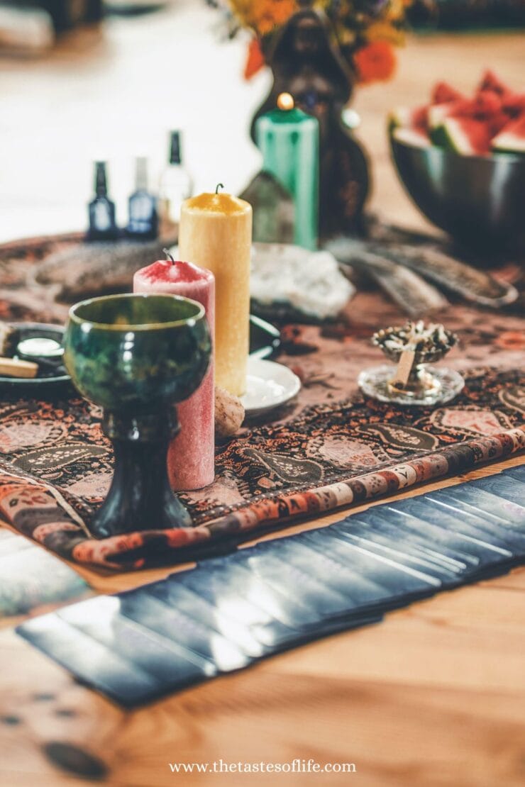 A decorative altar with candles, a chalice, crystals, tarot cards, and ritual items arranged on a patterned cloth, with flowers and a bowl of fruit in the background.