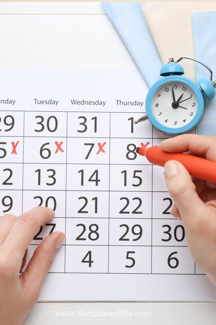 A hand marks a date on a calendar with a red pen; several dates are already crossed out. An analog blue alarm clock and pastel papers are placed nearby on a white surface.