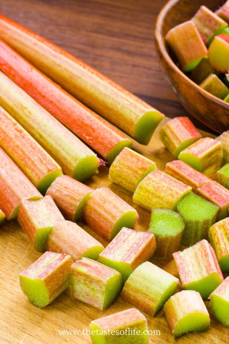 Fresh rhubarb stalks, some whole and some chopped into pieces, are arranged on a wooden cutting board. A wooden bowl filled with chopped rhubarb is visible in the background.