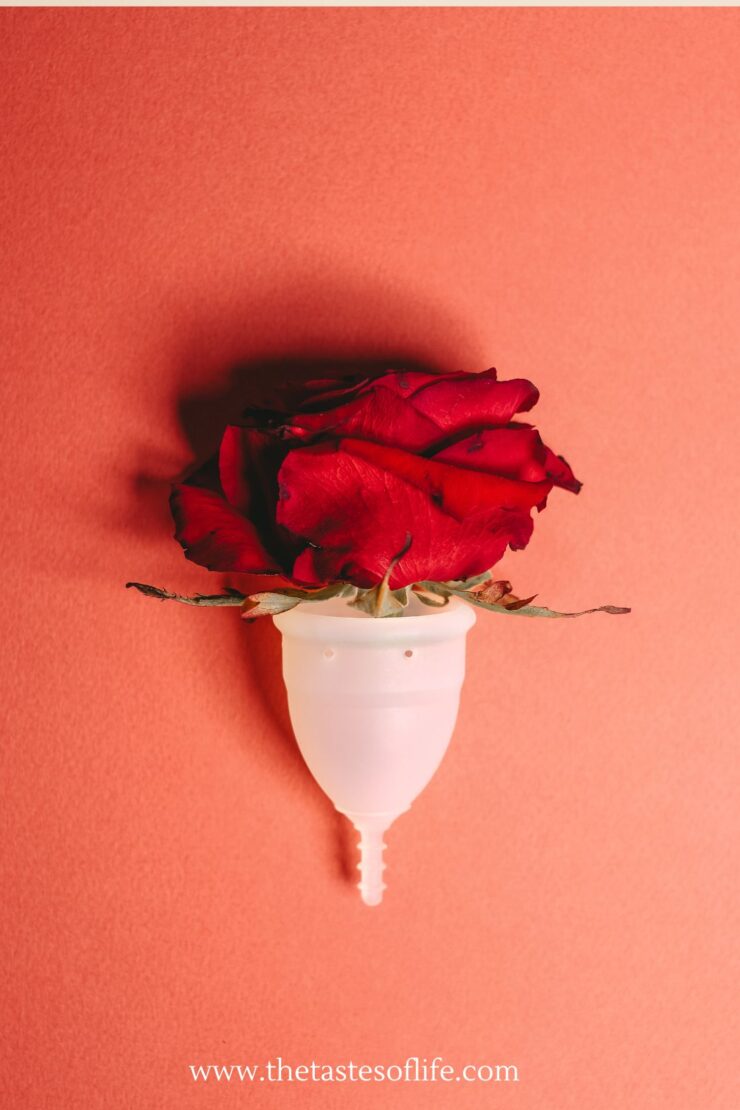 A red rose placed inside a white menstrual cup against a coral pink background, with the website www.thetastesoflife.com at the bottom.
