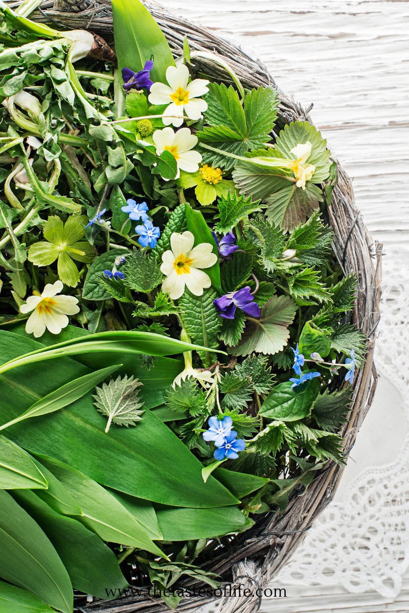 A woven basket filled with assorted fresh green herbs and colorful wildflowers, including yellow, blue, and purple blossoms, displayed on a white wooden surface.